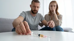 Мужчина сидит на диване и тянется за шприцом, рядом сидит шокированная женщина